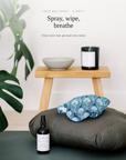 A yoga mat spray bottle and a blue patterned hot water pack resting on a dark gray cushion, with a small wooden stool, bowl, candle, and a large green plant leaf in the background. Text at the top reads 'YOGA MAT SPRAY - CLARITY' and 'Spray, wipe, breathe'.