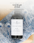 A bottle of Spritz Wellness London Yoga Mat Spray centered on a blue patterned fabric background, with soft beige wood tones around the edges and promotional text above reading 'Give the gift of ritual' and 'Yoga mat spray and a matching eye pillow.'.