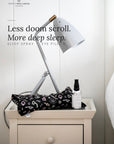 A bedside scene with a white nightstand holding a black eye pillow with white skull-and-crossbones pattern, a white desk lamp, and a small white bottle labeled Sleep Spray. Text on the image reads 'Less doom scroll.