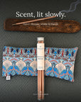 A tube of Spritz Wellness Moroccan Rose incense with two patterned eye pillows and a lit incense stick on a wooden tray