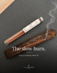 A tube of Spritz Wellness Moroccan Rose incense on a dark textured mat beside a wooden holder with a lit incense stick