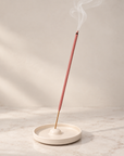 A pink incense stick stands upright in a round cream ceramic holder with white smoke rising against a soft beige background
