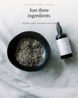 A yoga mat spray bottle next to a black bowl filled with dried lavender and tea tree leaves, set on a light gray textured fabric background with text overlay reading 'Just three ingredients'.