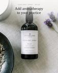 A bottle of Spritzwellness London Yoga Mat Spray in the 'Relax' scent, styled in a flat-lay arrangement with lavender flowers, a bowl of dried herbs, and a white mug on a textured white linen surface.