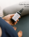 A hand holds a small spray bottle labeled Yoga Mat Spray Relax Calm the mind & body, with a blurred indoor setting in the background and text at the top.