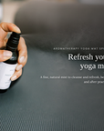 A hand sprays a fine mist from an aromatherapy yoga mat spray bottle onto a dark textured yoga mat, with white text on the right reading 'Refresh your yoga mat' and 'Aromatherapy Yoga Mat Spray'.