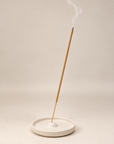 A single incense stick stands upright in a shallow round white ceramic dish, with a wisp of smoke curling upward against a plain warm beige studio background.