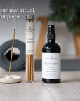 A product bundle from Spritz Wellness London including a Palo Santo incense stick in a glass tube, a black spray bottle of Yoga Mat Spray, and a white ceramic incense holder with a burning incense stick, arranged on a light wood surface with a blurred gray mat and green plant in the background.