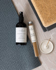 A styled flat-lay of a Lightwellness London Yogo Mat Spray bottle and a tube of Palo Santo incense sticks on a gray textured yoga mat, next to a white incense holder, on a light wood background.