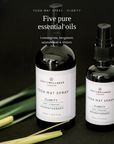 Two black glass bottles of Spritzwellness London essential oils on a dark background, with one bottle labeled 'Yoga Mat Spray Clarity' and the other partially visible. White text at the top reads 'Five pure essential oils' and lists the ingredients.