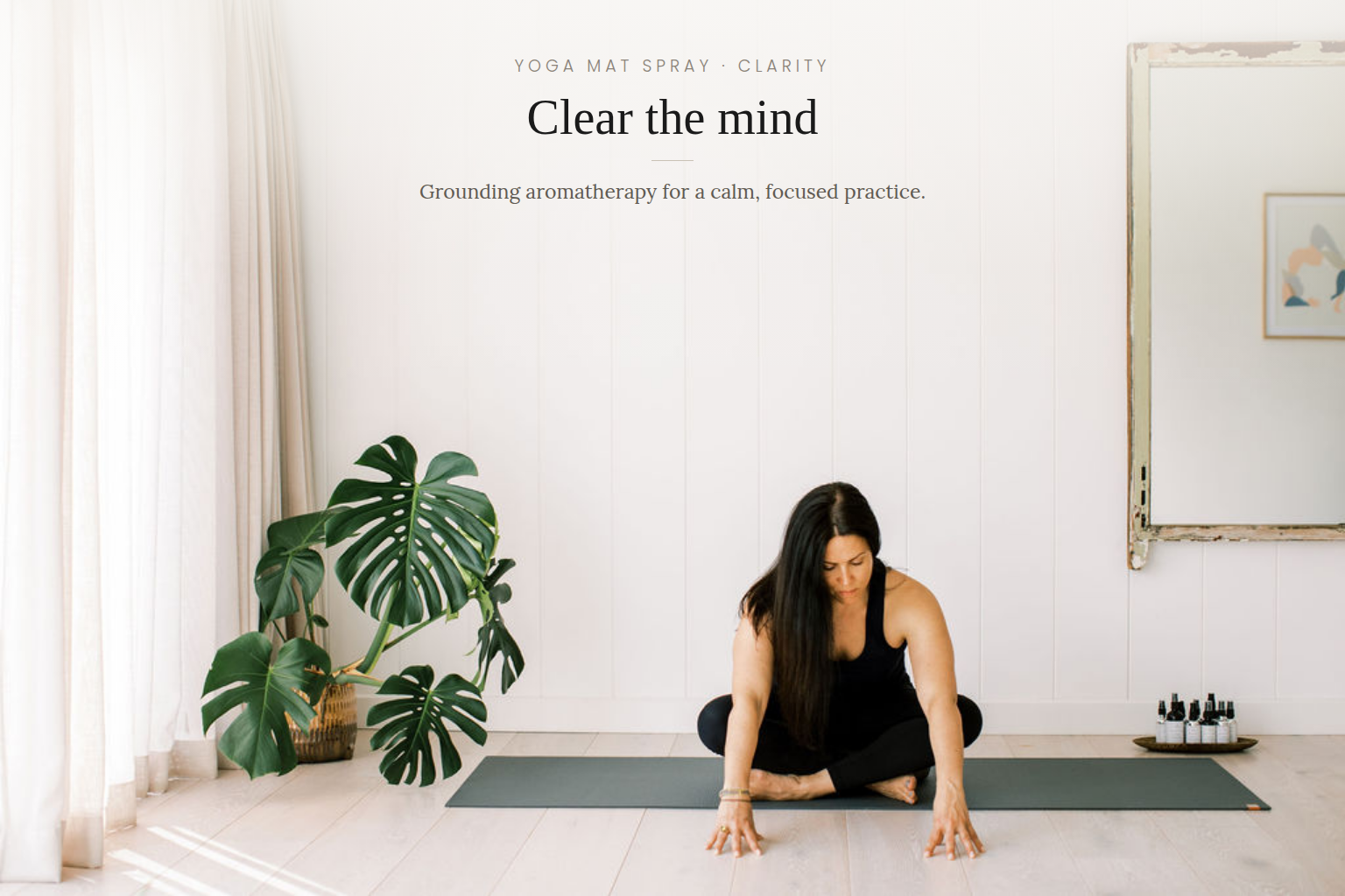 A woman in a black outfit sits cross-legged on a dark yoga mat in a bright, minimal room with white paneled walls, a large potted monstera plant, and a small tray of bottles. Text at the top reads 'YOGA MAT SPRAY - CLARITY' and 'Clear the mind'.