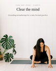A woman in a black outfit sits cross-legged on a dark yoga mat in a bright, minimal room with white paneled walls, a large potted monstera plant, and a small tray of bottles. Text at the top reads 'YOGA MAT SPRAY - CLARITY' and 'Clear the mind'.