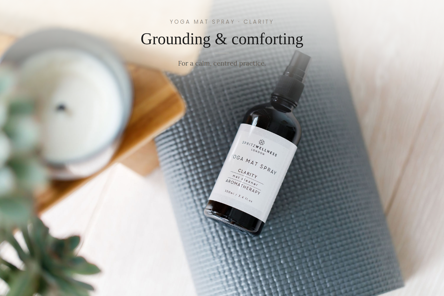 A bottle of Yoga Mat Spray by Groundness on a gray textured yoga mat, styled with a blurred candle and plant in a bright, light wood setting. Text overlay reads 'Grounding & comforting'.