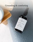 A bottle of Yoga Mat Spray by Groundness on a gray textured yoga mat, styled with a blurred candle and plant in a bright, light wood setting. Text overlay reads 'Grounding & comforting'.