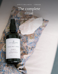 A bottle of Spritz Wellness London Yoga Mat Spray placed on a patterned blue and beige yoga mat next to a white crystal, with overlaid text reading 'The complete ritual' and 'Refresh your mat, then rest your eyes.'.