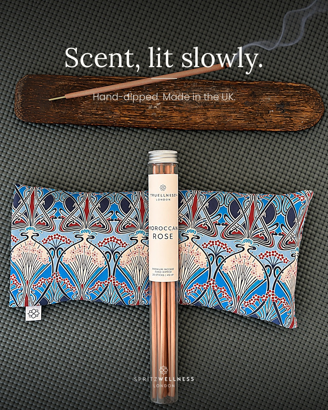 A tube of Spritz Wellness Moroccan Rose incense with two patterned eye pillows and a lit incense stick on a wooden tray