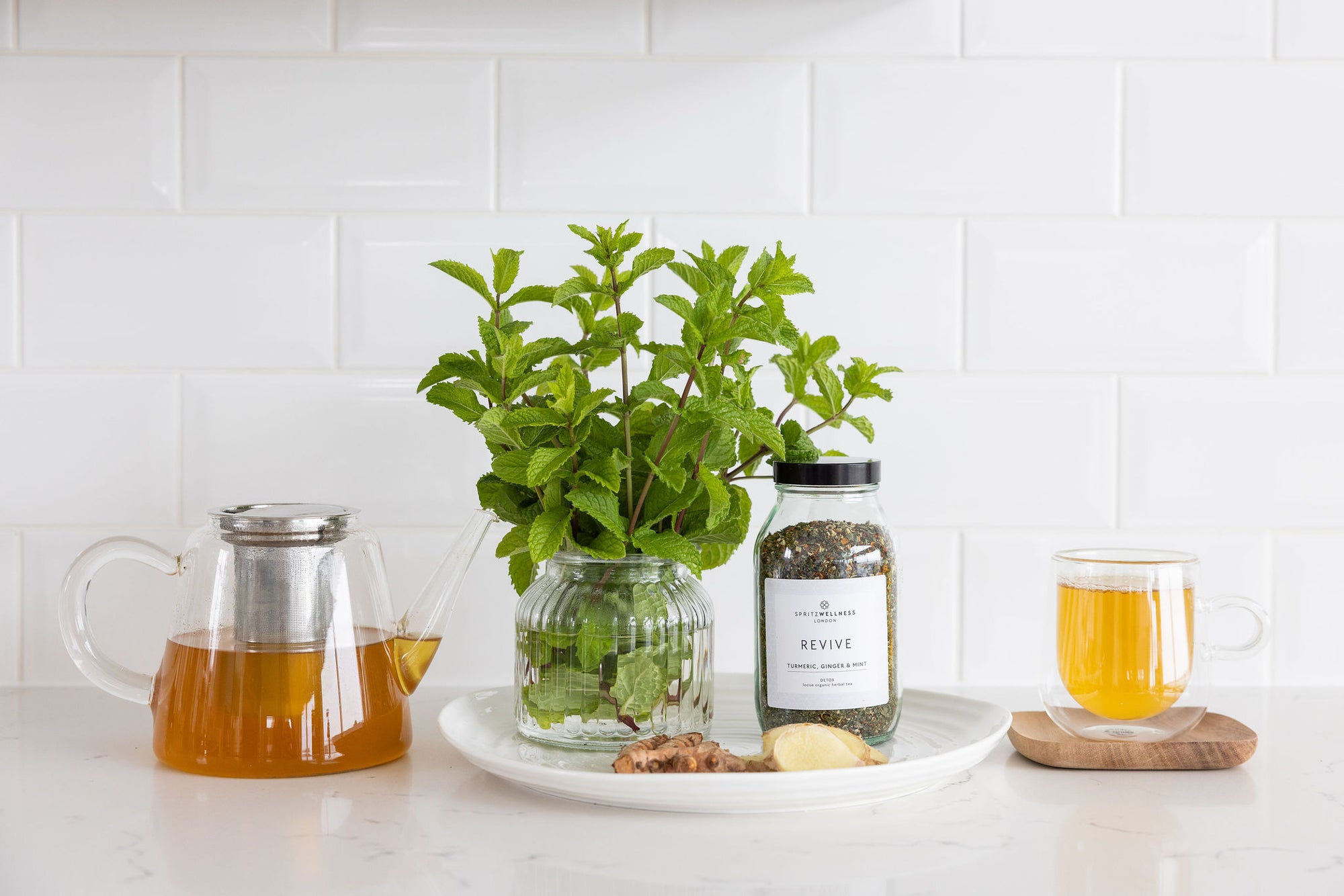 A lifestyle photo showing Spritz Wellness Revive tea ingredients: a jar of loose leaf tea, fresh mint, ginger, and a glass pot of steeped golden tea.