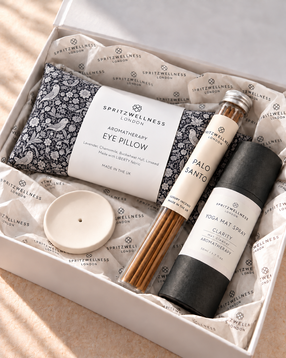 A gift box from Spritzwellness London containing an aromatherapy eye pillow, a Palo Santo incense stick, a yoga mat spray bottle, and a small white candle, all arranged on branded tissue paper.