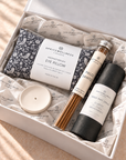 A gift box from Spritzwellness London containing an aromatherapy eye pillow, a Palo Santo incense stick, a yoga mat spray bottle, and a small white candle, all arranged on branded tissue paper.
