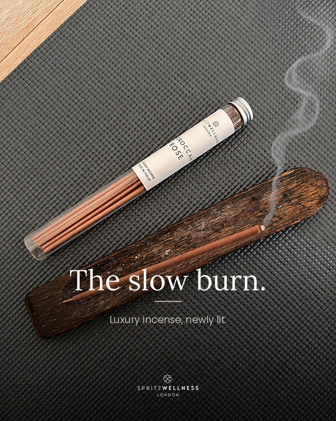 A tube of Spritz Wellness Moroccan Rose incense on a dark textured mat beside a wooden holder with a lit incense stick
