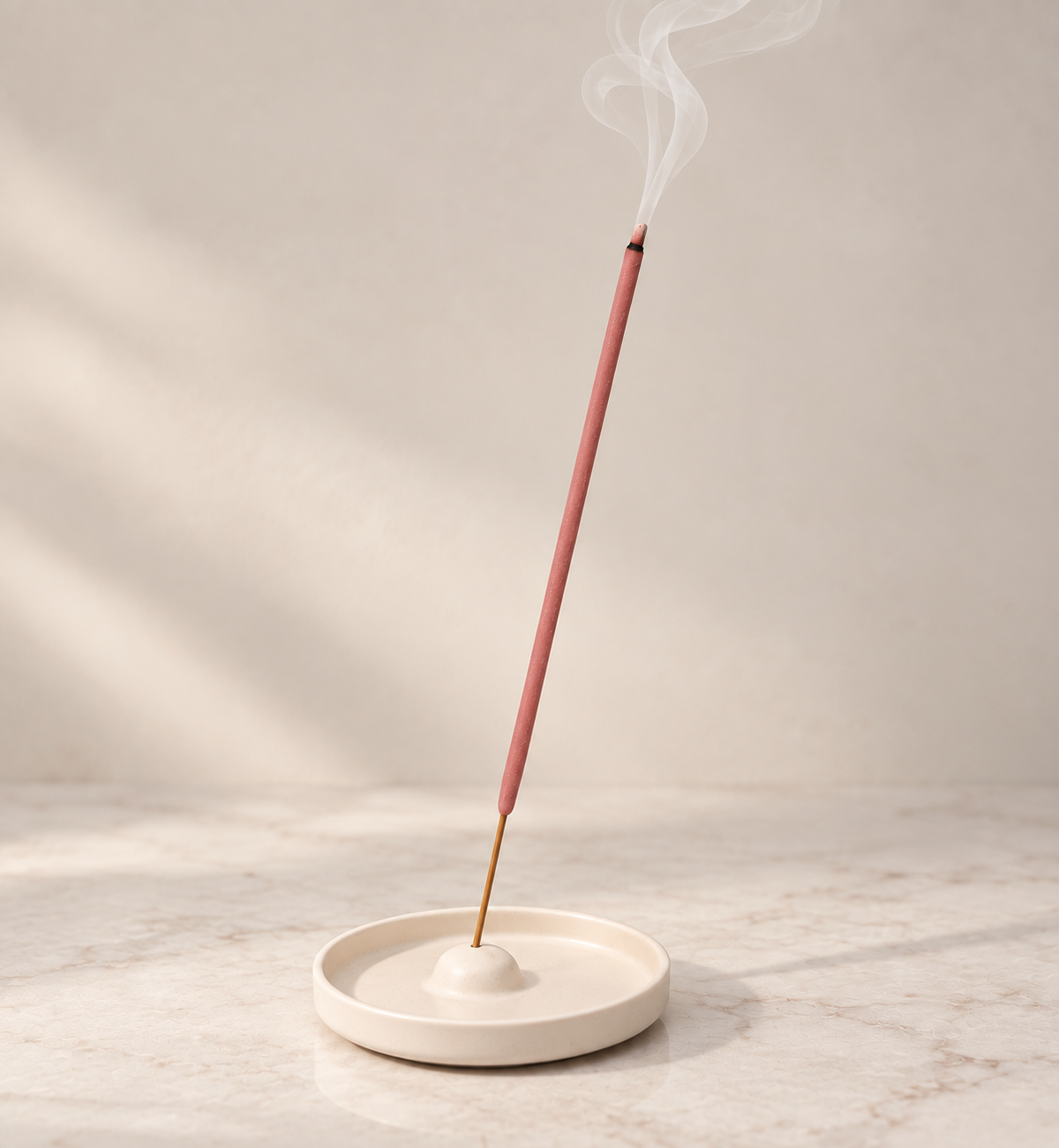 A pink incense stick stands upright in a round cream ceramic holder with white smoke rising against a soft beige background