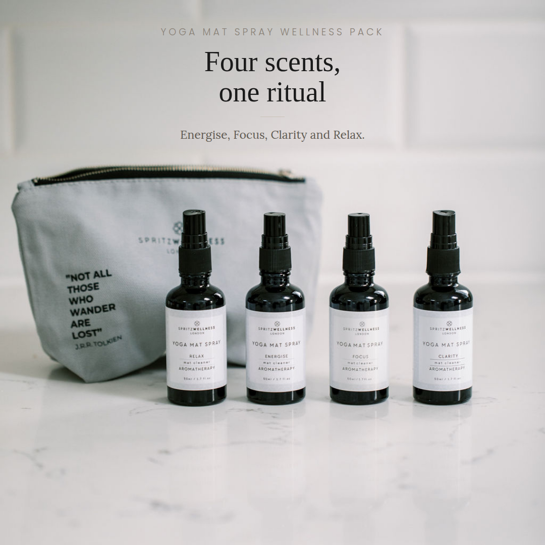 A yoga mat spray wellness pack featuring four small black spray bottles with white labels lined up on a white marble surface next to a gray zippered pouch. Text above reads 'Four scents, one ritual' and 'Yoga Mat Spray Wellness Pack'.