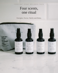 A yoga mat spray wellness pack featuring four small black spray bottles with white labels lined up on a white marble surface next to a gray zippered pouch. Text above reads 'Four scents, one ritual' and 'Yoga Mat Spray Wellness Pack'.