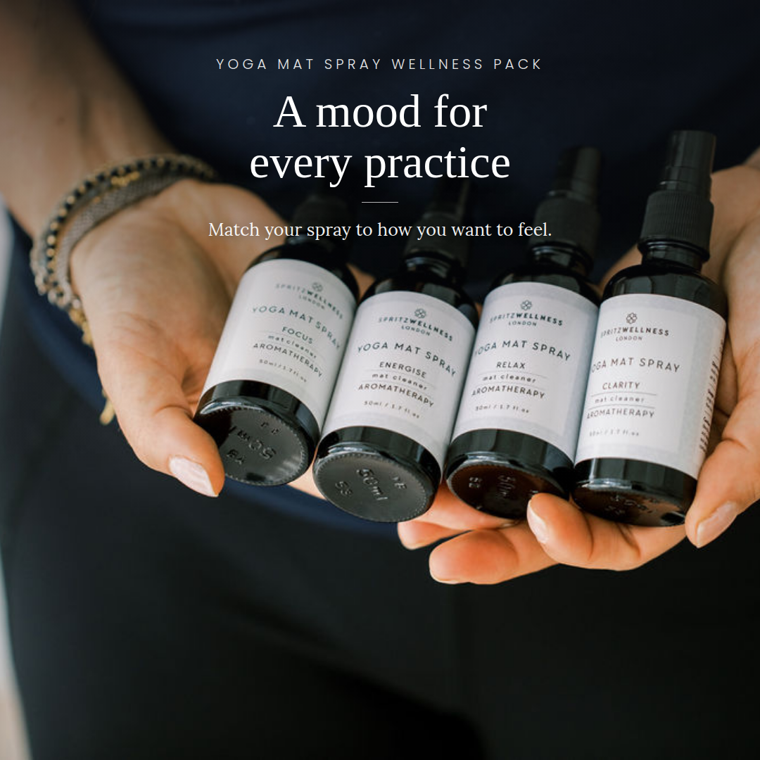 Four small yoga mat spray bottles held in two hands against a dark navy shirt background, with overlaid text reading 'YOGA MAT SPRAY WELLNESS PACK' and 'A mood for every practice'.