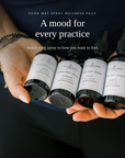 Four small yoga mat spray bottles held in two hands against a dark navy shirt background, with overlaid text reading 'YOGA MAT SPRAY WELLNESS PACK' and 'A mood for every practice'.