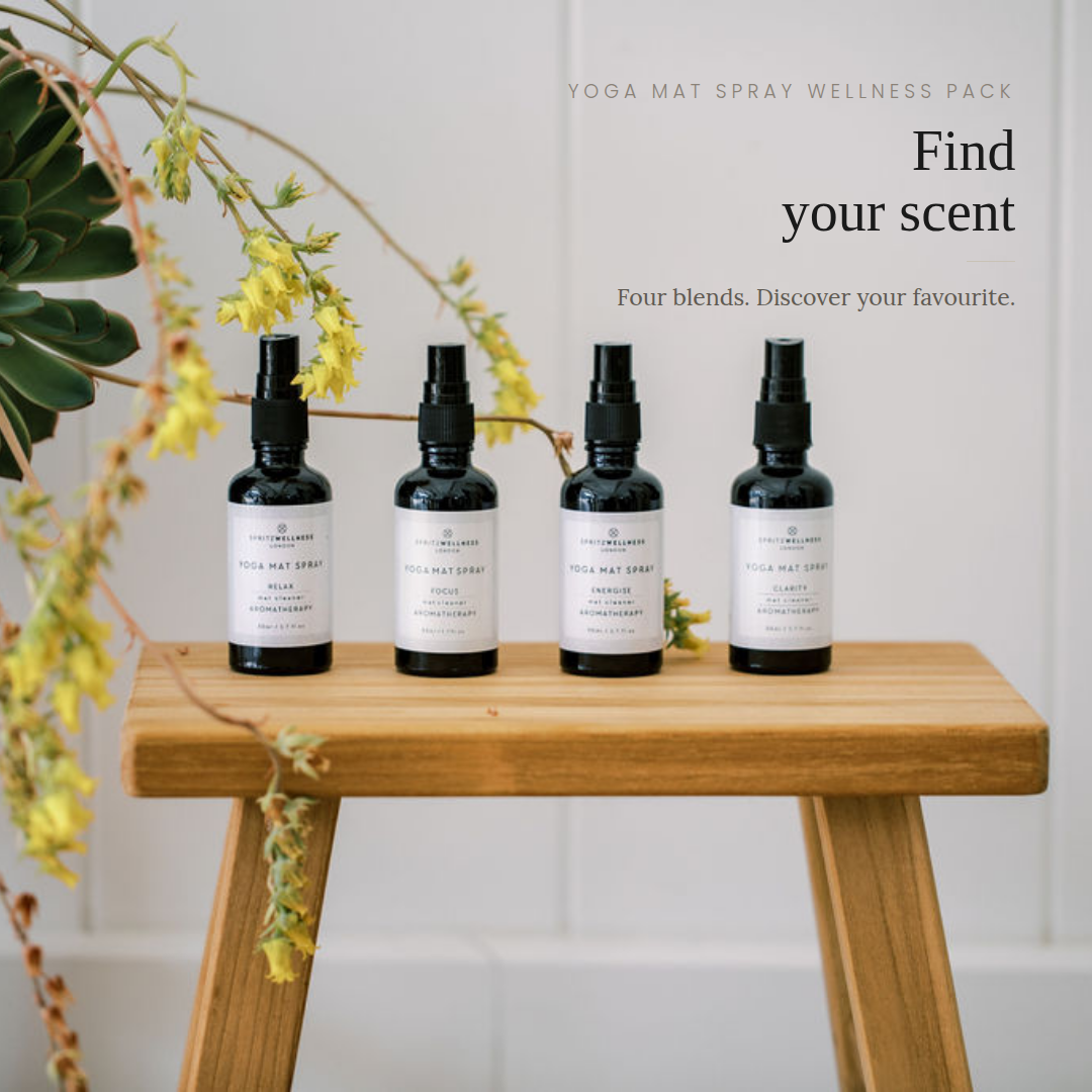 Four black yoga mat spray bottles with white labels are lined up on a wooden stool against a white paneled wall, with decorative green leaves and yellow flowers hanging from the left side. Text on the right reads 'YOGA MAT SPRAY WELLNESS PACK', 'Find your scent', and 'Four blends.