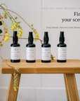 Four black yoga mat spray bottles with white labels are lined up on a wooden stool against a white paneled wall, with decorative green leaves and yellow flowers hanging from the left side. Text on the right reads 'YOGA MAT SPRAY WELLNESS PACK', 'Find your scent', and 'Four blends.