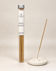 A tube of Hitzwell London Palo Santo incense sticks stands next to a small white ceramic incense holder on a warm beige background.