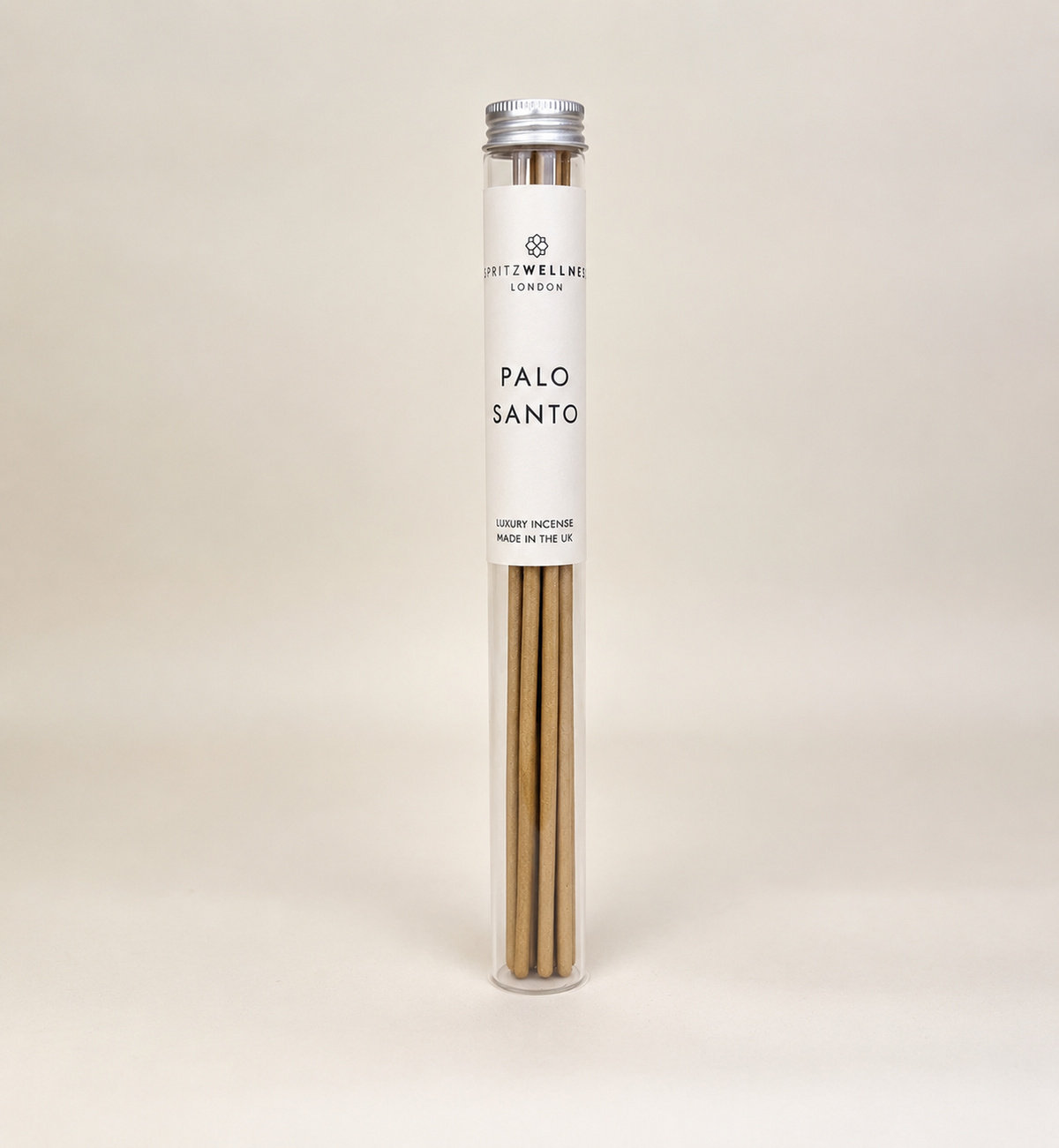 A tall, clear cylindrical tube containing Palo Santo incense sticks, sealed with a silver cap and wrapped in a white paper label that reads 'spritz wellness' and 'PALO SANTO'. The product is photographed on a seamless, warm off-white background.