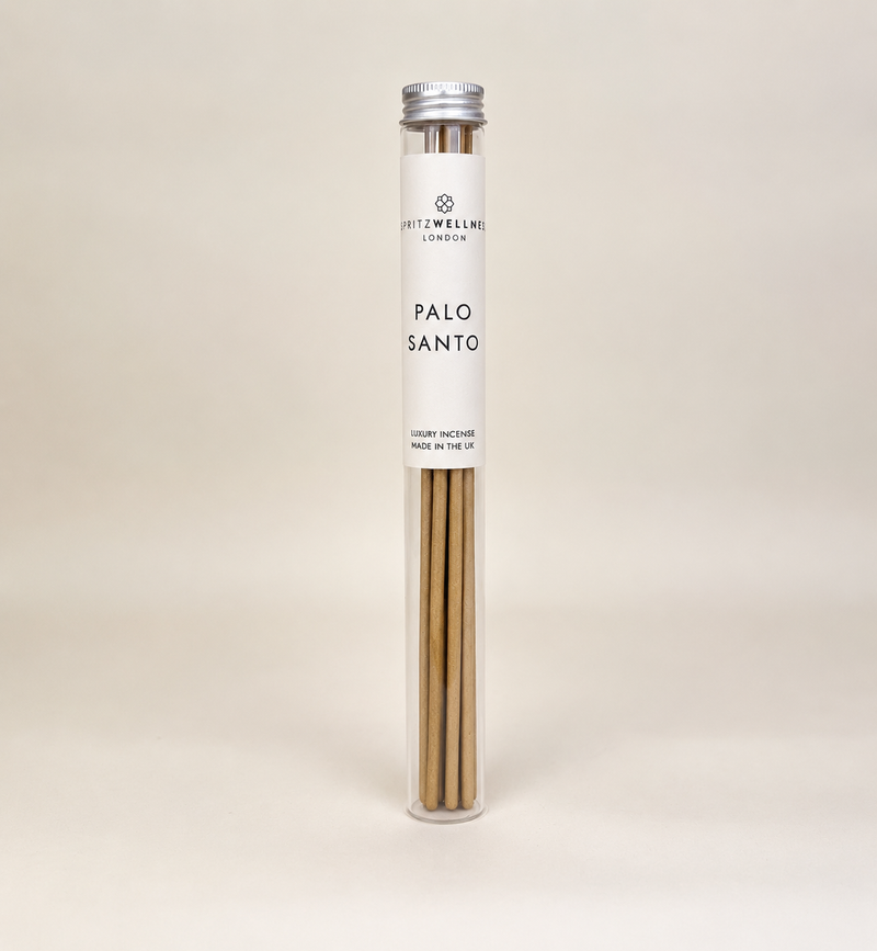A tall, clear cylindrical tube containing Palo Santo incense sticks, sealed with a silver cap and wrapped in a white paper label that reads 'spritz wellness' and 'PALO SANTO'. The product is photographed on a seamless, warm off-white background.