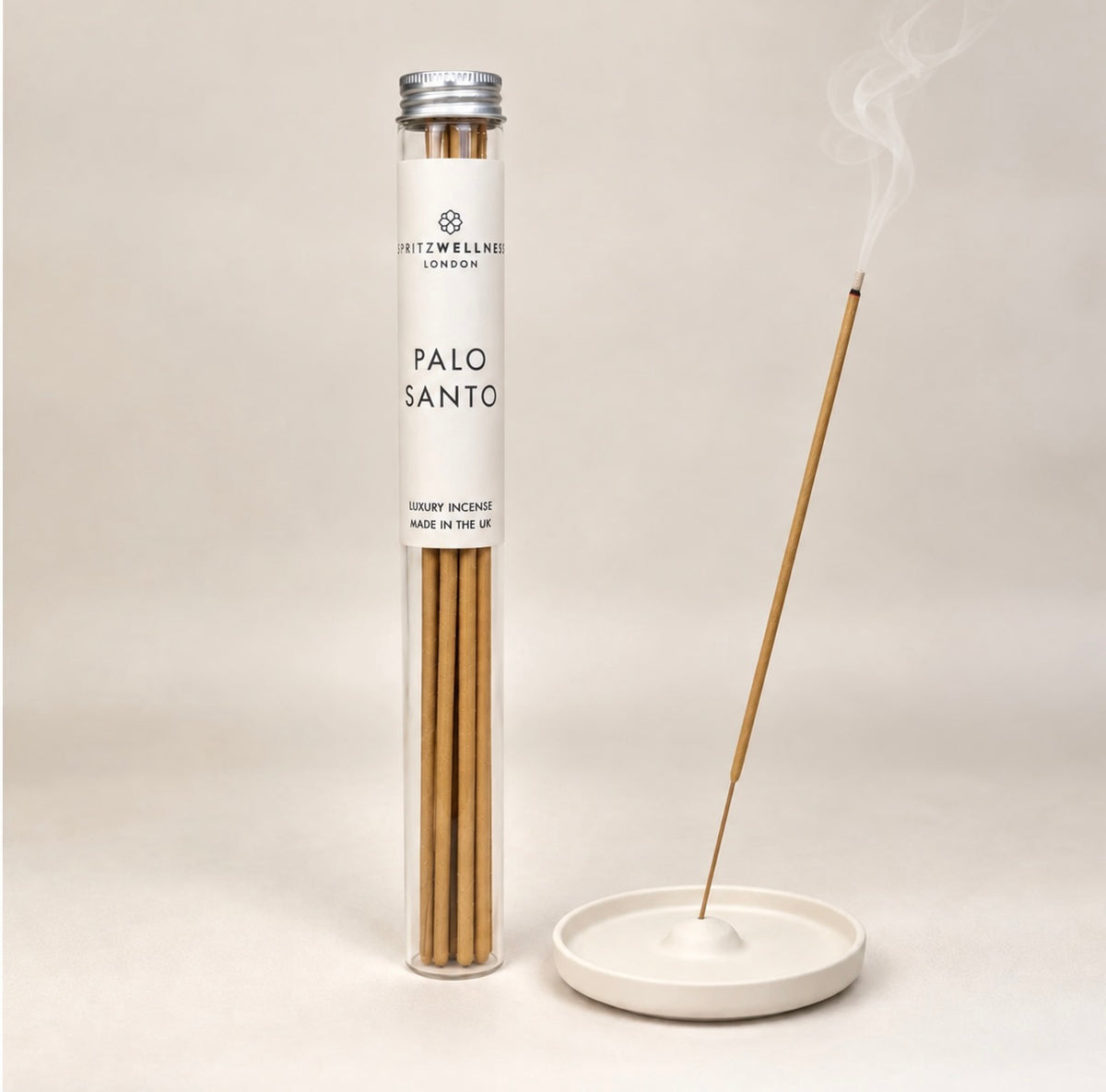 A tube of Spritz Wellness London Palo Santo incense sticks stands beside a white ceramic incense holder with one stick burning and smoke rising, on a light beige background.