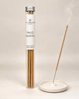 A tube of Spritz Wellness London Palo Santo incense sticks stands beside a white ceramic incense holder with one stick burning and smoke rising, on a light beige background.