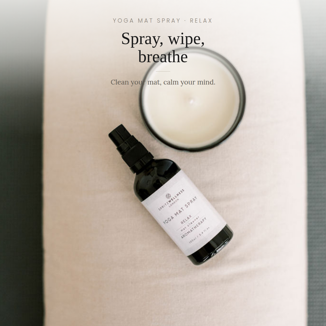 A bottle of Yogi Mat Spray by Serenity Studios lies on a cream-colored yoga mat next to an open candle jar, with text overlay reading 'YOGA MAT SPRAY - RELAX' and 'Spray, wipe, breathe'.