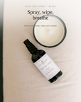 A bottle of Yogi Mat Spray by Serenity Studios lies on a cream-colored yoga mat next to an open candle jar, with text overlay reading 'YOGA MAT SPRAY - RELAX' and 'Spray, wipe, breathe'.