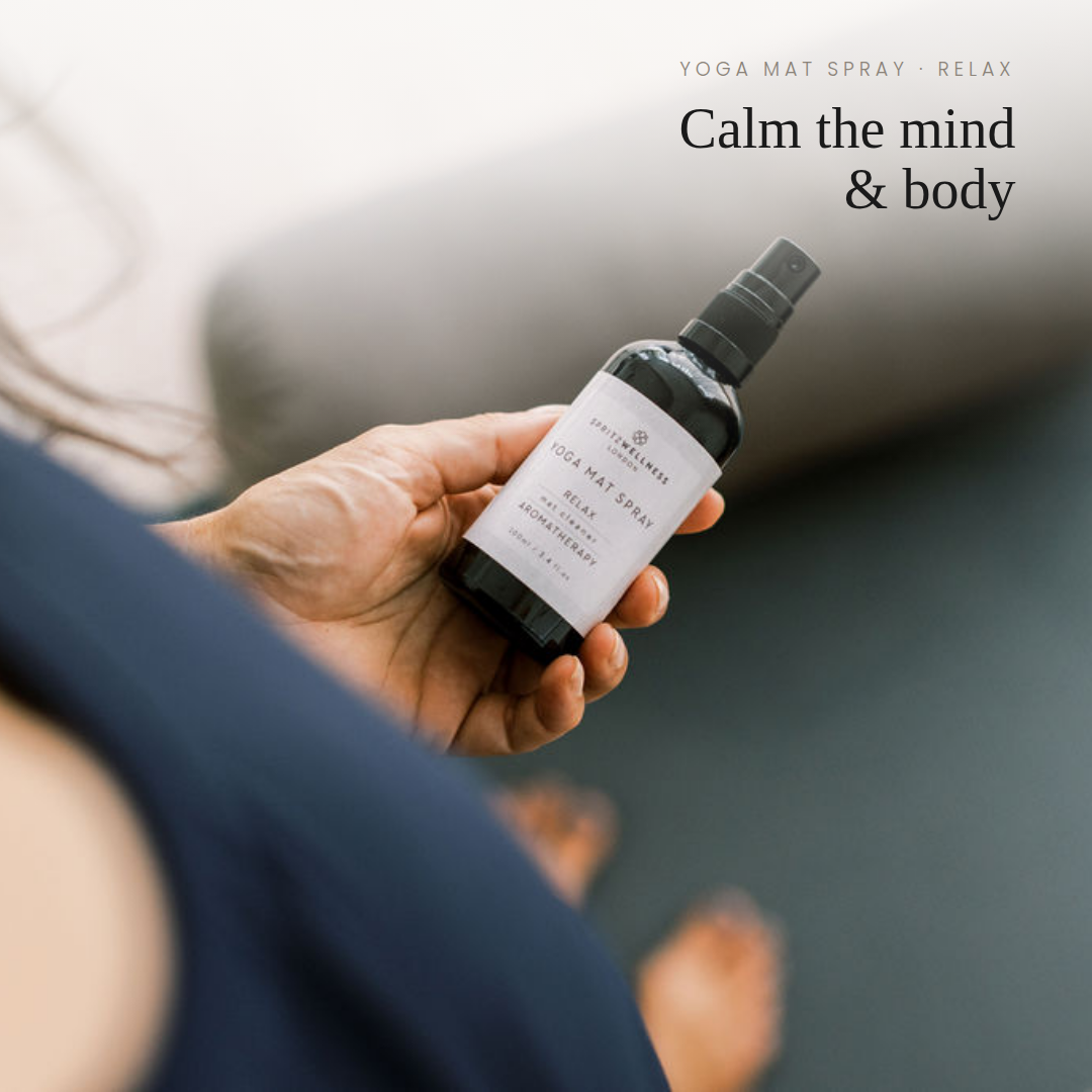 A hand holds a small spray bottle labeled Yoga Mat Spray Relax Calm the mind & body, with a blurred indoor setting in the background and text at the top.
