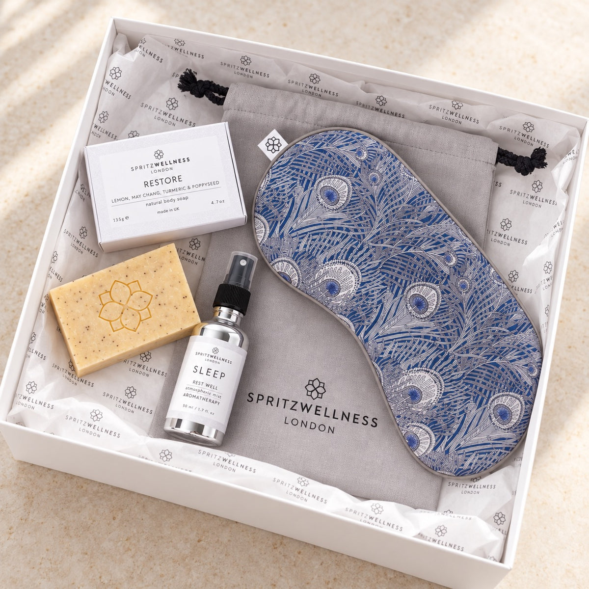A curated self-care gift set from Spritzwellness London arranged in a white box. The set includes a blue and white patterned sleep mask, a gray drawstring pouch, a small white spray bottle labeled Sleep, a beige bar of soap, and a white box labeled Restore.
