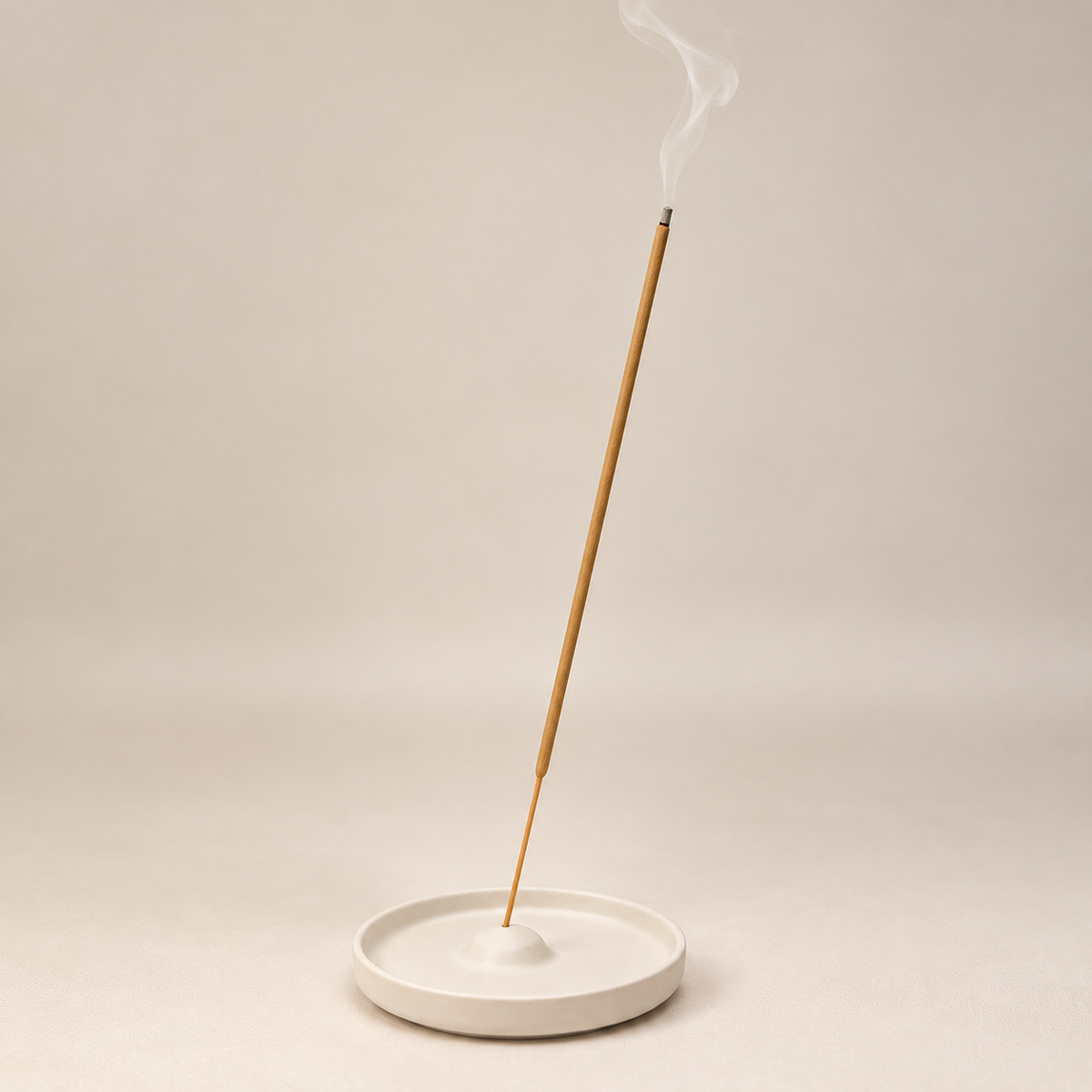 A single incense stick stands upright in a shallow round white ceramic dish, with a wisp of smoke curling upward against a plain warm beige studio background.