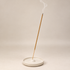 A single incense stick stands upright in a shallow round white ceramic dish, with a wisp of smoke curling upward against a plain warm beige studio background.