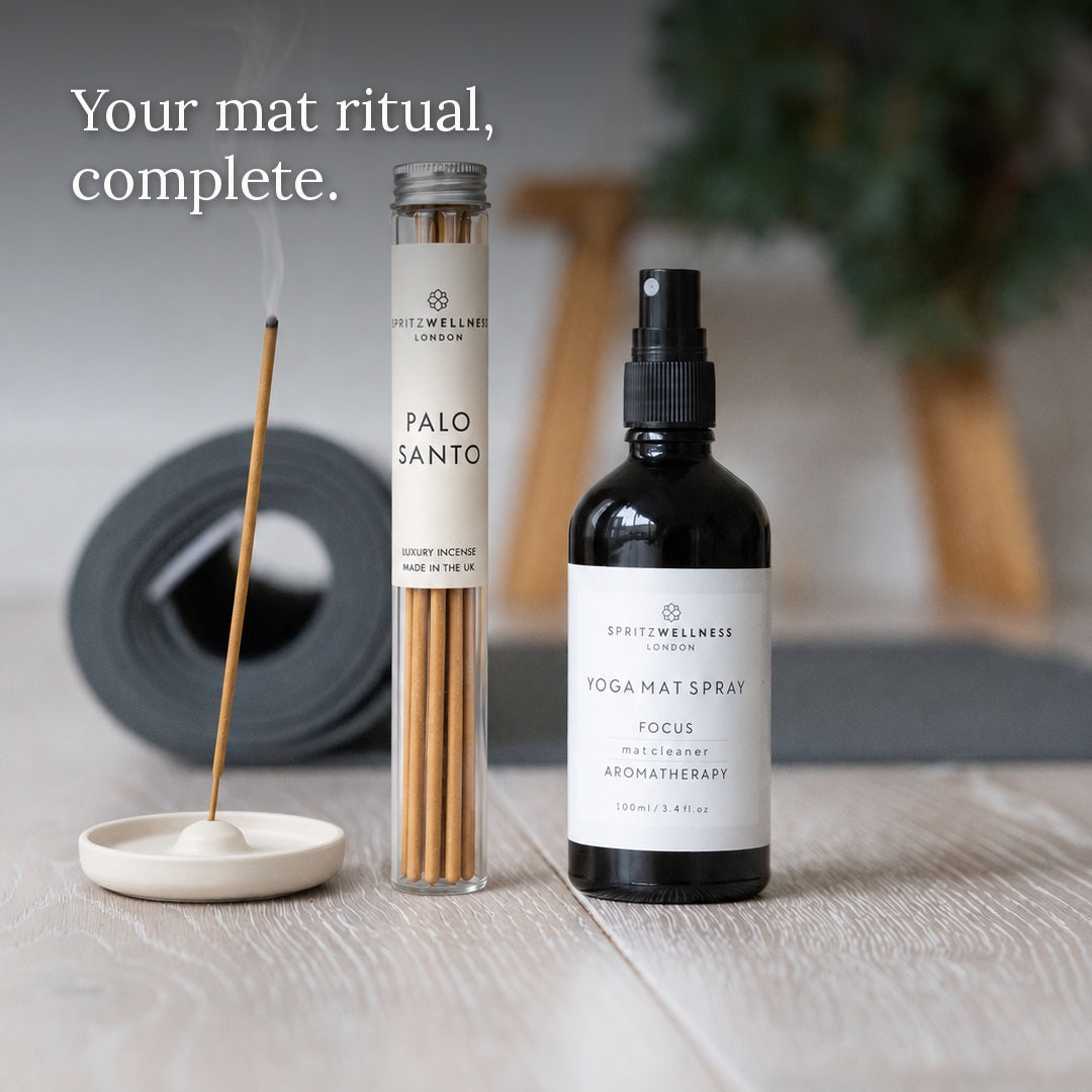 A product bundle from Spritz Wellness London including a Palo Santo incense stick in a glass tube, a black spray bottle of Yoga Mat Spray, and a white ceramic incense holder with a burning incense stick, arranged on a light wood surface with a blurred gray mat and green plant in the background.