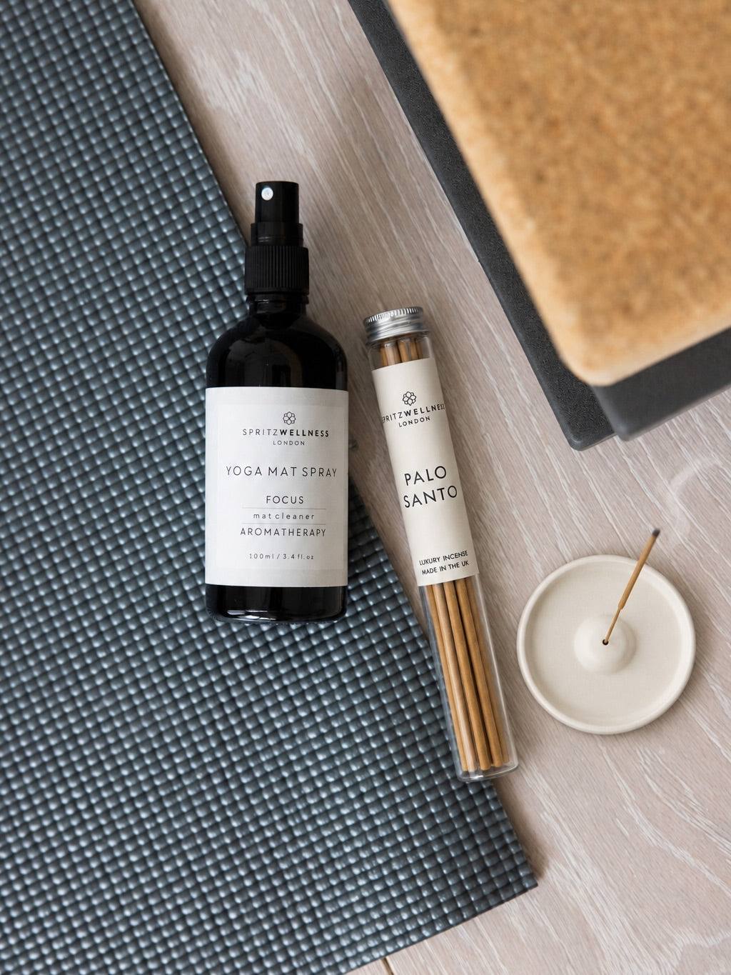 A styled flat-lay of a Lightwellness London Yogo Mat Spray bottle and a tube of Palo Santo incense sticks on a gray textured yoga mat, next to a white incense holder, on a light wood background.